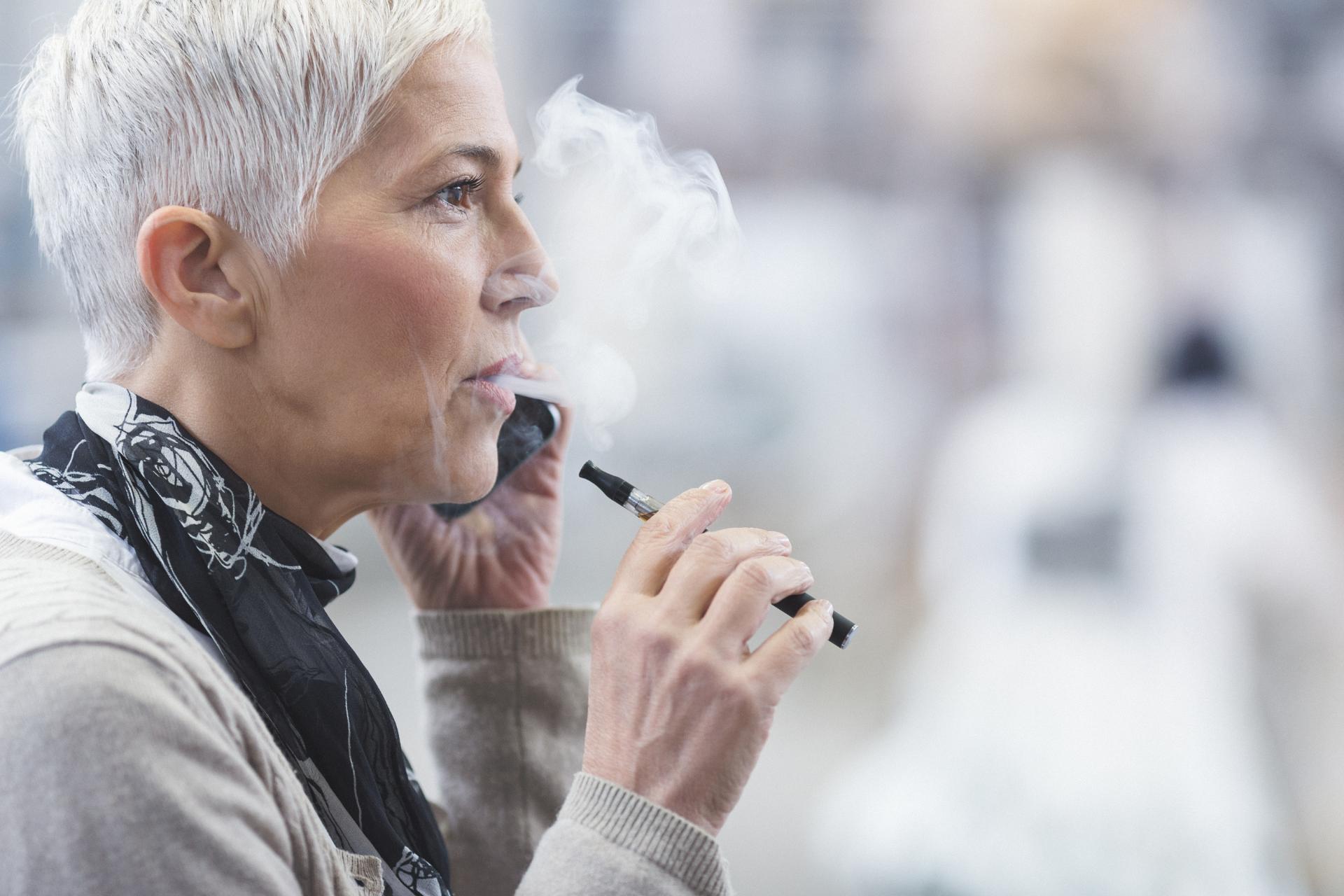 Berry Good Vibes E-Liquid, One Love E-Liquid. Older woman with short gray hair vaping and talking on a smartphone, with smoke visible in the air.