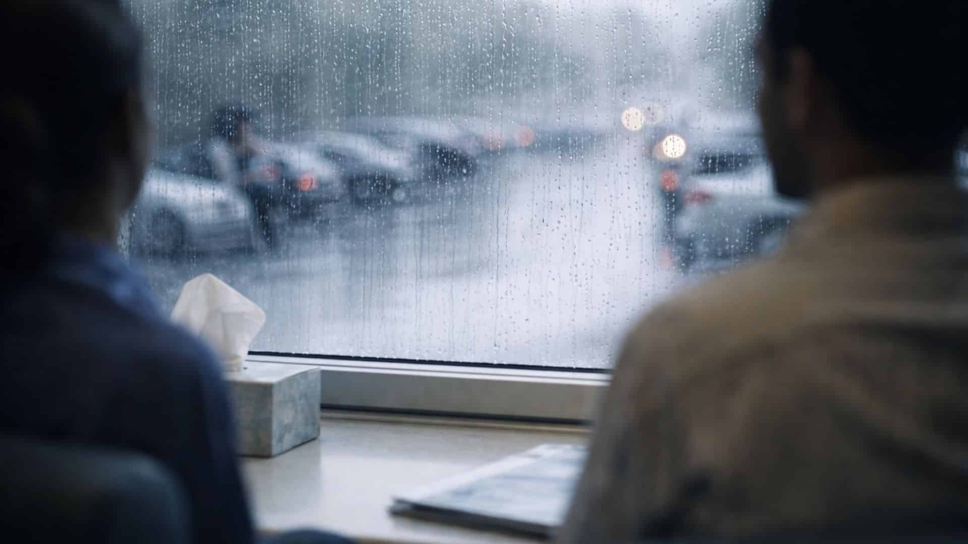 A man and woman sit by a rainy window, blurred cars visible outside. A tissue box and papers are on the table, creating a contemplative mood.